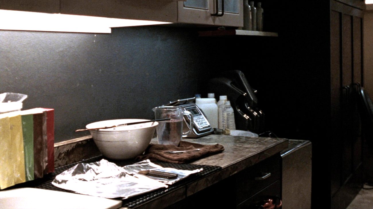 A salon colour station on a dark worktop: a plastic tint bowl with a brush laid across it, a glass measuring jug, a digital scale, developer bottles, sheets of foil laid out flat and a folded towel, under a strip light.