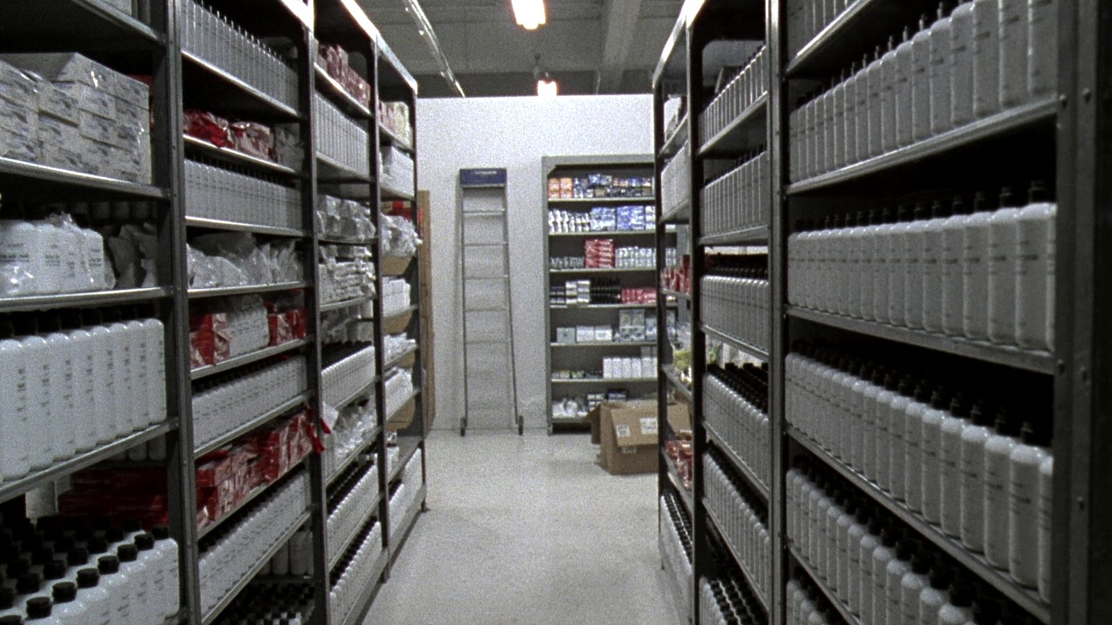 An aisle inside a professional beauty supply stockroom: grey steel shelving racks holding rows of identical white and dark bottles, cardboard cases stacked at the end of the aisle, a rolling ladder, concrete floor under fluorescent light.